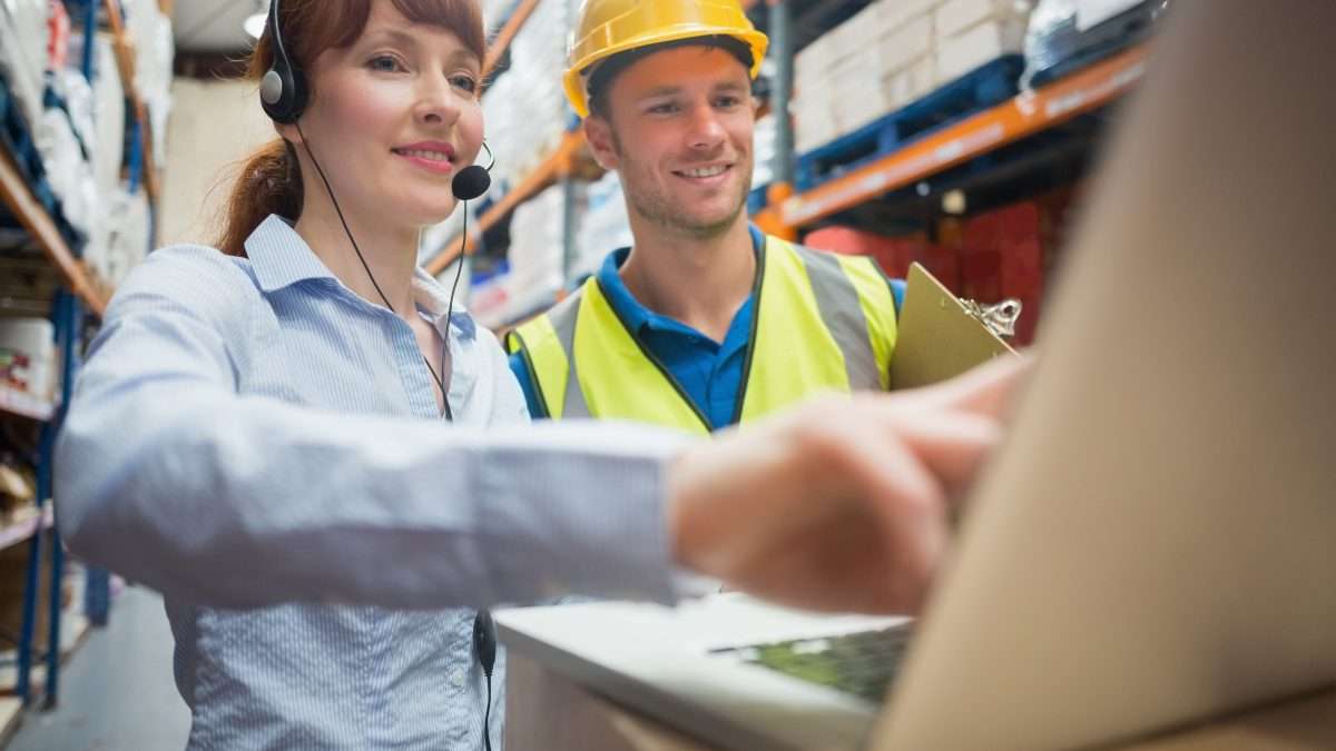 A supervisor works on a computer with a work placement student wearing a hardhat and safety vest in a warehouse