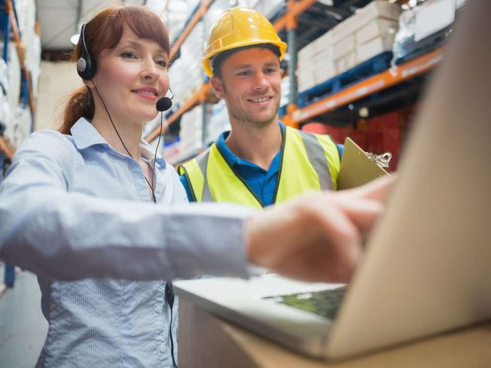 Un superviseur travaille sur un ordinateur aux côtés d’un stagiaire qui porte un casque de protection et un gilet de sécurité dans un entrepôt