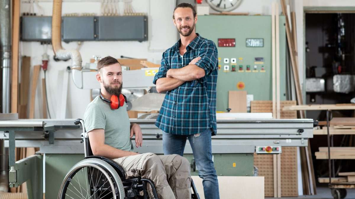 A worker in a wheelchair and his colleague are seen in a carpenter's workshop.