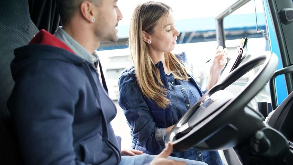 A female trucking logistics worker checks something on a clipboard and speaks with a male driver while she stands in the open door of his truck cab.