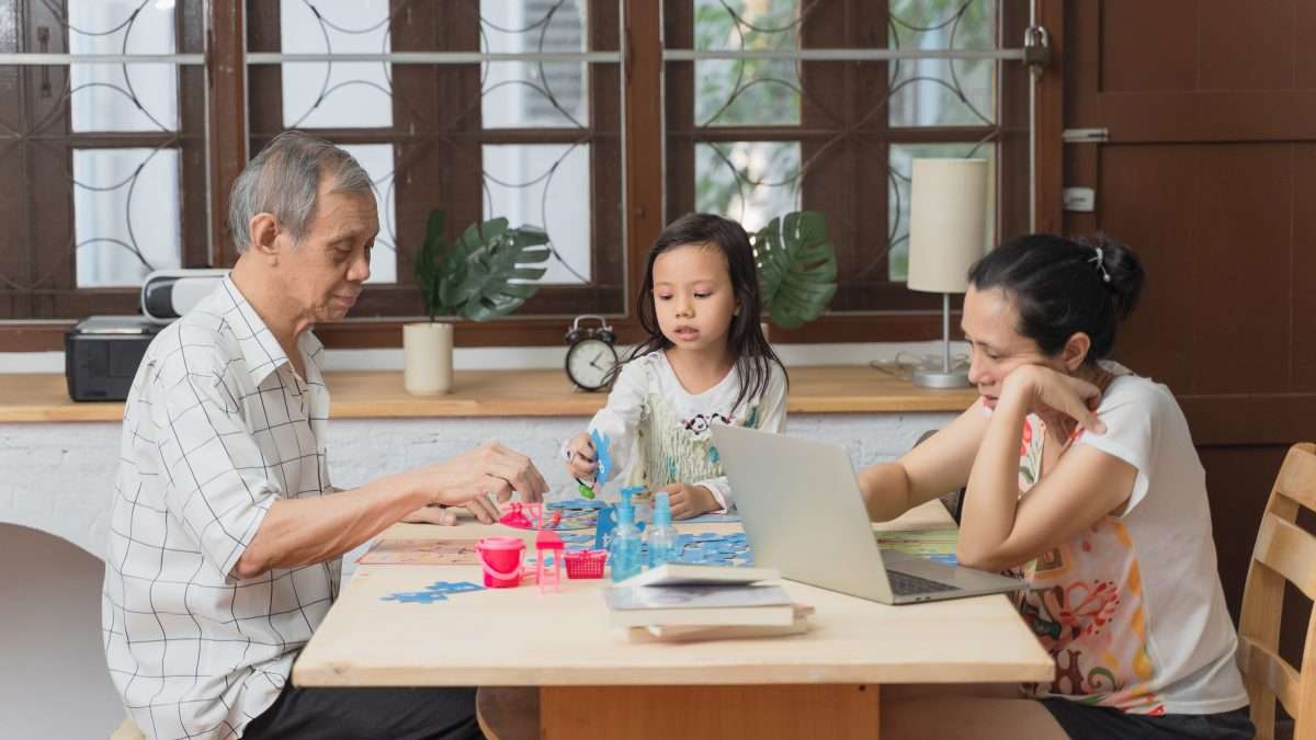 Woman working with laptop while daughter does homework with grandfather.