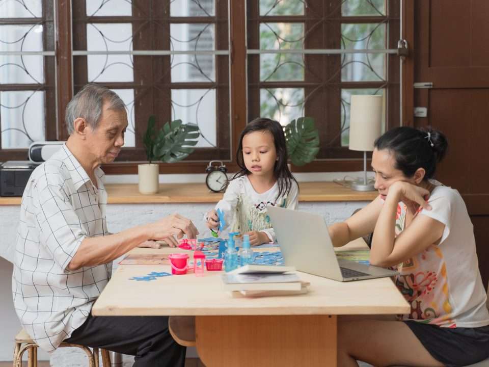 Woman working with laptop while daughter does homework with grandfather.