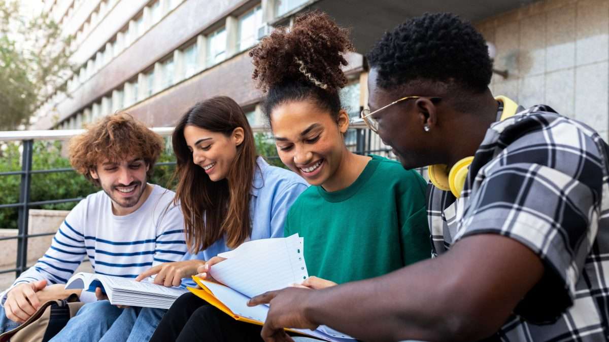 A group of post-secondary students smile and chat while sitting on the steps outside a campus building.