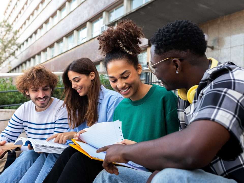 A group of post-secondary students smile and chat while sitting on the steps outside a campus building.