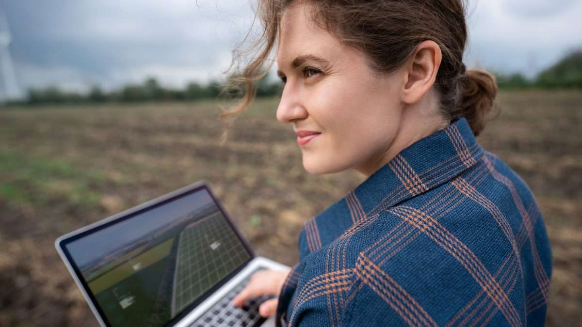 A woman holds a laptop while surveying a farm field with a large wind turbine standing at its far edge.