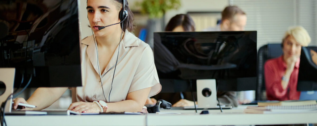 Young adult woman with disability working in call center, wearing headset and using computer in team office. Customer support and inclusive workplace concept for service business marketing
