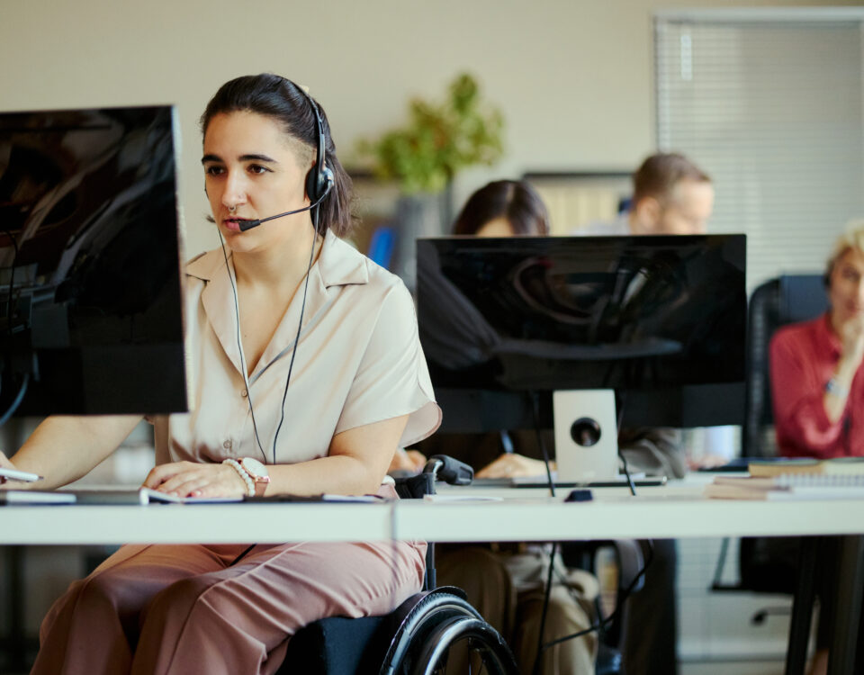 Young adult woman with disability working in call center, wearing headset and using computer in team office. Customer support and inclusive workplace concept for service business marketing