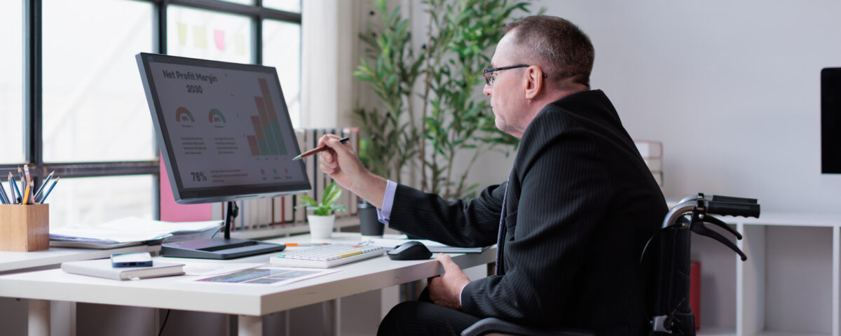 Business professional in wheelchair analyzing financial data on computer monitor, working in modern office, showing diversity and inclusion in workforce
